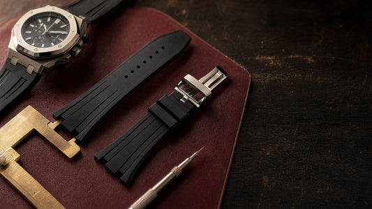 A black rubber watch strap with a steel folding clasp on a burgundy leather mat next to a brass caliper, a steel sports watch and a spring bar tool.