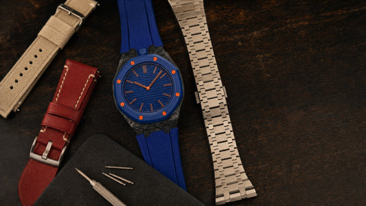 A blue forged carbon Royal Pop style watch on a dark workbench surrounded by a sand canvas strap, a burgundy leather strap, a steel link bracelet and spring bars.