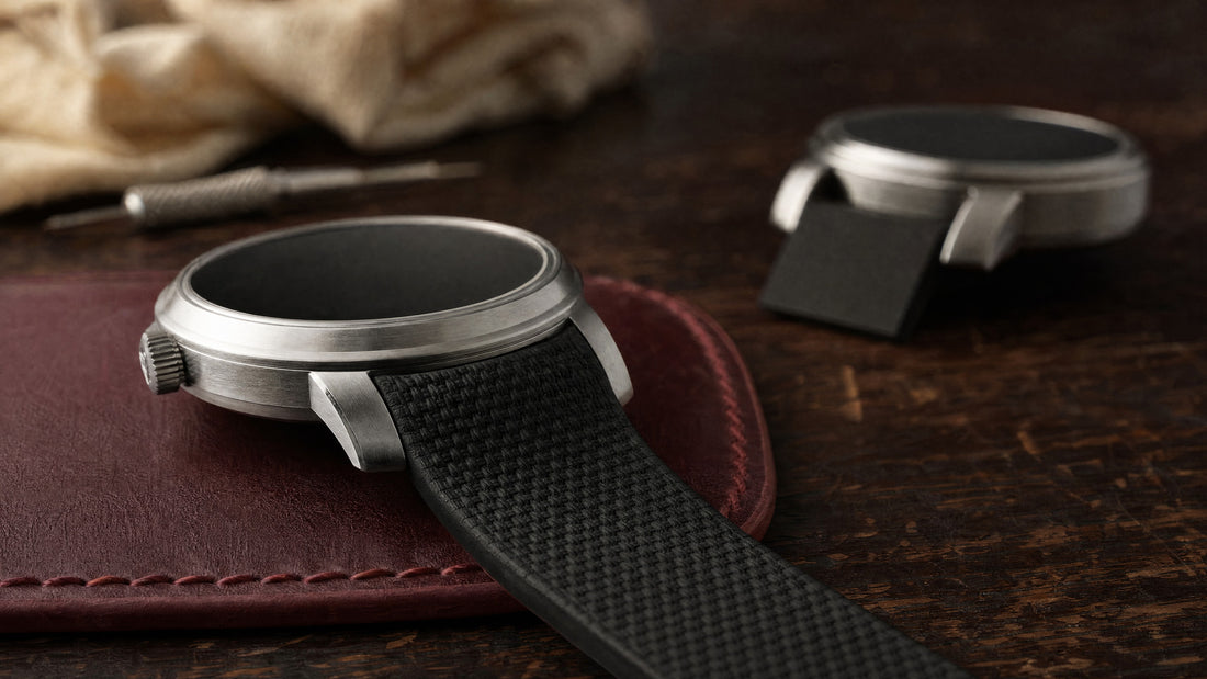 Low-angle macro of a round steel watch with a black textured rubber strap whose curved end fills the gap between the lugs, with a straight-end strap and visible gap out of focus behind.