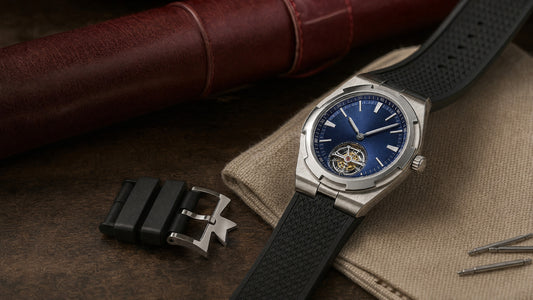 A steel Vacheron Constantin Overseas watch with a blue sunburst dial on a black textured rubber strap, next to a steel Maltese cross buckle, spring bars and a sand canvas cloth.