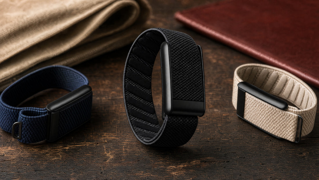 Three woven WHOOP bands in black, navy and sand with matte black sensor pods on a dark wooden workbench.